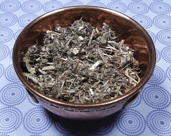 May include: A copper bowl filled with dried, light brown and gray herbs. The bowl is sitting on a purple and white patterned surface.