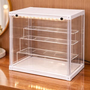 May include: A white display cabinet with clear acrylic sides and shelves. The cabinet has an LED light strip along the top. The unit is designed to showcase collectibles or cosmetics. The dimensions are in centimetres.