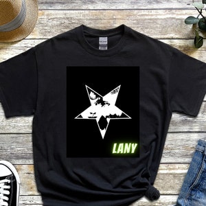 May include: Black t-shirt featuring a white star design with a detailed graphic inside. The word "LANY" is printed in green below the star. The shirt is made of a soft material.