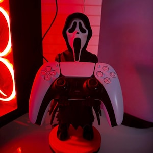 Può includere: Una figura di Ghostface in bianco e nero che tiene in mano un controller PlayStation 5 bianco e nero. La figura è su una base rossa. Lo sfondo include una console PlayStation 5 e illuminazione rossa.