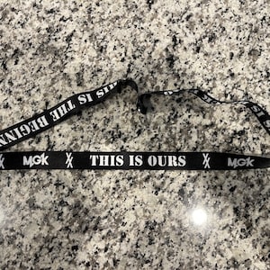 Puede incluir: Lanyard negro con texto blanco que dice "This is Ours" y "MGK" a cada lado. El texto "This is the beginning" est&aacute; impreso en la parte posterior del lanyard.