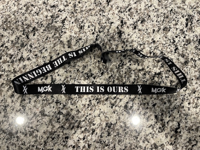 Puede incluir: Lanyard negro con texto blanco que dice "This is Ours" y "MGK" a cada lado. El texto "This is the beginning" est&aacute; impreso en la parte posterior del lanyard.