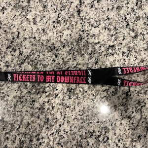 Puede incluir: Lanyard negro con texto rosa que dice "X TICKETS TO MY DOWNFALL X".