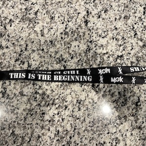 Puede incluir: Lanyard negro con texto blanco que dice "This is the beginning" y "MGK" repetido dos veces. El lanyard tiene un cierre de metal plateado.