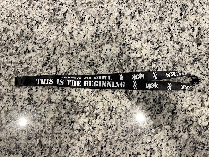 Puede incluir: Lanyard negro con texto blanco que dice "This is the beginning" y "MGK" repetido dos veces. El lanyard tiene un cierre de metal plateado.