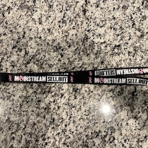Puede incluir: Lanyard negro con texto blanco que dice "Mainstream Sellout" con un s&iacute;mbolo de anarqu&iacute;a rosa. El texto se repite en ambos lados del lanyard.