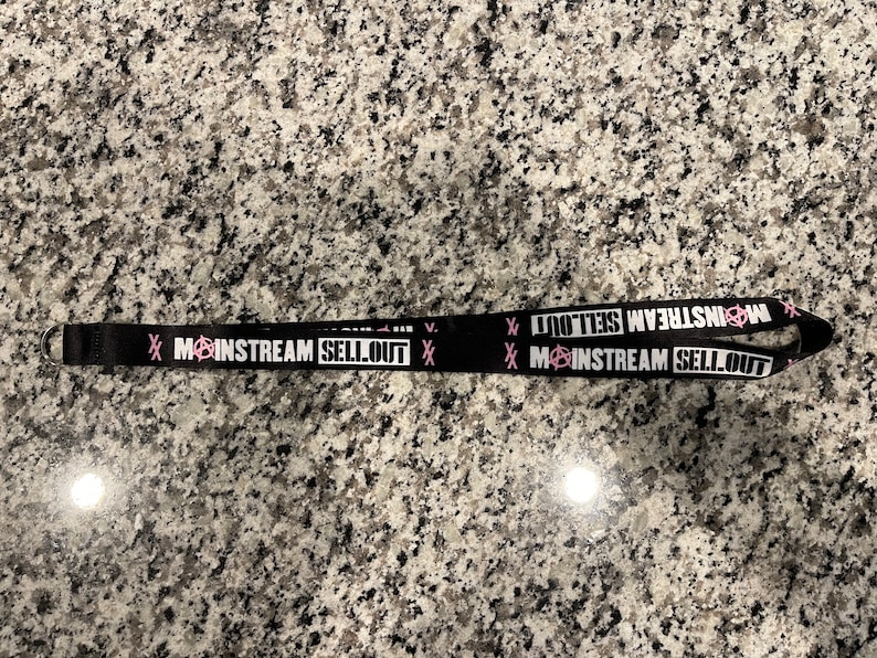 Puede incluir: Lanyard negro con texto blanco que dice "Mainstream Sellout" con un s&iacute;mbolo de anarqu&iacute;a rosa. El texto se repite en ambos lados del lanyard.
