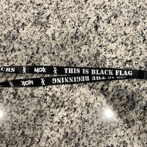 Puede incluir: Lanyard negro con texto blanco que dice "This is Black Flag - The Beginning" y "MGK" con una X a cada lado.