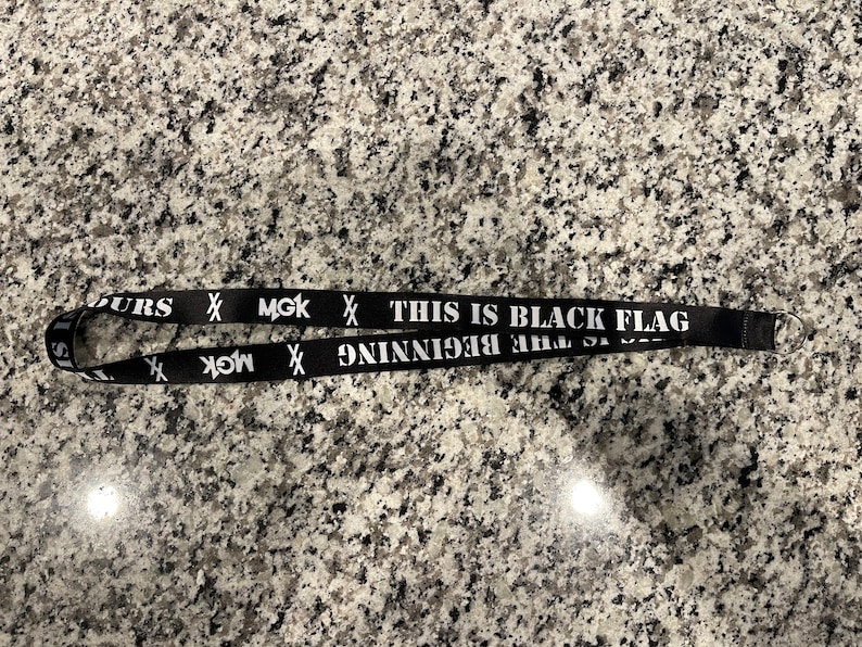 Puede incluir: Lanyard negro con texto blanco que dice "This is Black Flag - The Beginning" y "MGK" con una X a cada lado.