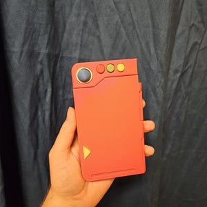 Pode incluir: Um dispositivo portátil vermelho com um botão azul, três botões menores e um triângulo amarelo. O dispositivo é segurado na mão de uma pessoa contra um fundo azul escuro. O dispositivo parece ser um console de jogos ou um acessório.