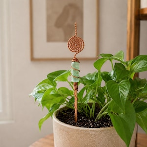 May include: A copper wire plant stake featuring a spiral top and a cylindrical light green stone accent. The wire is wrapped around the stone and extends into a straight stem, designed to be inserted into the soil of a potted houseplant.