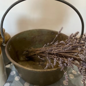 May include: A vintage brass cauldron with a dark metal handle, containing dried lavender. Two ceramic jars with cork and ceramic lids are in the background. The cauldron has a rustic, aged appearance.