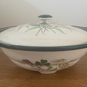 May include: A white ceramic serving bowl with a lid. The bowl and lid are decorated with a floral pattern in green, purple, and yellow. The rim and handle are a muted green colour. The bowl is sitting on a wooden surface.