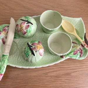 May include: A green ceramic serving tray with floral designs, featuring four small bowls and a spoon. The tray is decorated with a leafy pattern and has a small knife.