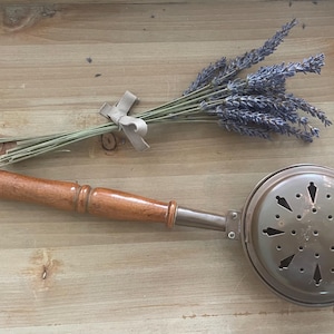 May include: A bunch of dried lavender tied with a pale ribbon, alongside a vintage metal and wood bed warmer. The bed warmer features a wooden handle and a round, perforated metal head.