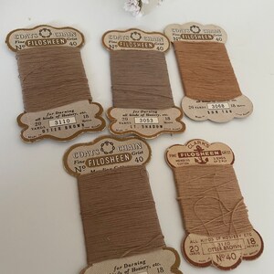 May include: Five spools of Coats & Clark's FiloSheen thread in various shades of brown. The spools are labelled with the thread colour, yardage, and the brand name. The spools are arranged in a row on a white surface.