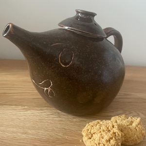 May include: A brown ceramic teapot with a lid and a spout. The teapot has a unique design with white markings on the side. The teapot is sitting on a wooden surface next to two light brown biscuits.