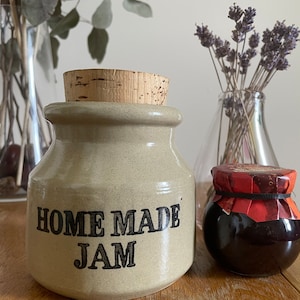 May include: A beige ceramic jar with a cork stopper and the words "HOME MADE JAM" in black. A small jar of jam with a red and brown label sits next to it. Dried lavender and eucalyptus are in glass vases.