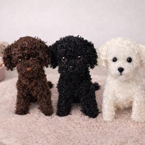 Könnte beinhalten: Drei Plüschhunde mit lockigem, strukturiertem Fell in Dunkelbraun, Schwarz und Weiß. Jedes Stofftier hat dunkle Augen, eine kleine Nase und Schlappohren und sitzt aufrecht in einer Reihe auf einer runden Matte mit weicher, flauschiger Oberfläche.