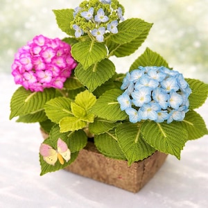Puede incluir: Un arreglo de plantas de ganchillo hecho a mano con vibrantes flores de hortensia rosas, celeste y azules. Las flores están en una maceta cuadrada marrón, con hojas verdes y una mariposa decorativa. Una encantadora decoración floral hecha a mano.