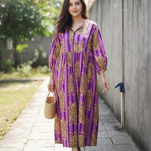 May include: A long, purple dress with a tiger stripe pattern. The dress has three-quarter sleeves and a V-neck. A small, woven handbag is held in the hand. The dress is worn barefoot on a stone walkway.