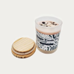 Op de afbeelding: Een romige, naar koffie geurende sojawas kaars in een matglazen pot met een houten deksel. De kaars heeft een lichtbruine kleur met accenten van koffiebonen. De pot heeft de tekst "Apple Candle Co."