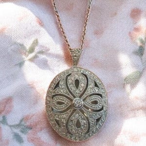 Könnte beinhalten: Eine zarte goldene Kettenkette mit einem runden, ovalen Anhänger. Der Anhänger ist aus Metall und hat ein florales Design mit kleinen, klaren Steinen.