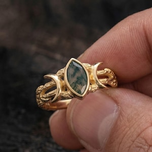 May include: A gold ring featuring a marquise-cut moss agate stone at the centre, flanked by two crescent moon shapes on either side. The band has a textured, branch-like appearance, and the metal has a polished, warm yellow gold finish.