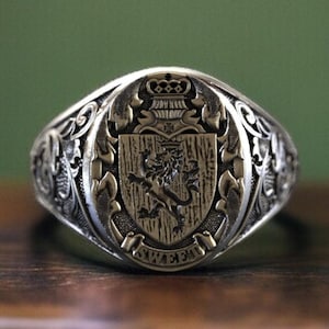 Op de afbeelding: Een zilveren zegelring met een ovale kop. De ring heeft een gedetailleerd wapen met een leeuw, een kroon en krullen. De ring ligt op een donker houten oppervlak met een groene achtergrond. Een klassiek sieraad.