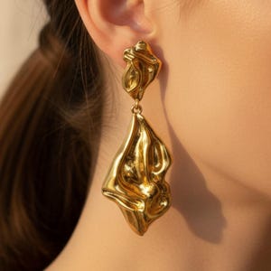 May include: Gold-coloured, abstract dangle earrings. The earrings have a unique, textured design with an organic, flowing shape. The earrings are shown on a person's ear, with a simple gold hoop earring visible.