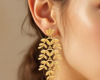 May include: Gold-colored leaf-shaped earrings. The earrings feature a floral design at the top, with multiple cascading leaf-like elements. The earrings are worn on a person's ear, with the background out of focus.