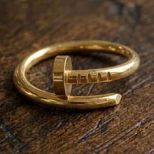 May include: A gold-colored ring with a nail-head design. The ring features a curved band with a nail head and a nail shaft detail. The ring is made of a shiny, metallic material and is set against a dark wooden surface.