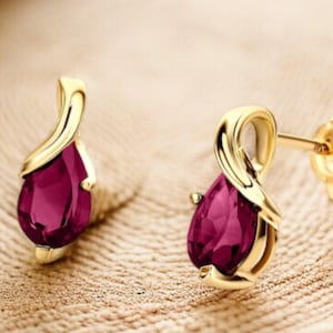 May include: A pair of gold-colored stud earrings with a teardrop-shaped, deep red gemstone. The earrings have a curved, decorative design above the gemstone. The earrings are displayed on a textured, light brown surface.