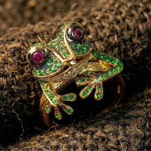 May include: A gold frog ring with green and red gemstones. The frog's eyes are red, and its body is covered in green stones. The ring is set on a textured brown surface.