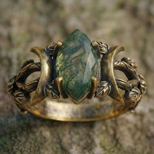 May include: A detailed close-up of a bronze-colored ring featuring a large, marquise-cut green gemstone. The ring has an ornate design with leaf and vine details, creating a vintage or fantasy aesthetic. The gemstone has a unique, moss-like pattern.