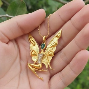 May include: Gold fairy pendant necklace. The pendant depicts a seated fairy with detailed wings, a green gemstone, and sparkling accents. The necklace is shown against a natural green backdrop.