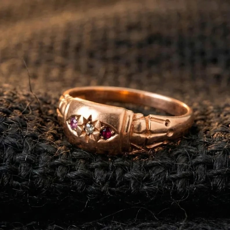 May include: A gold ring with three inset gemstones. The center stone is a clear diamond, flanked by a pink and a red gemstone. The ring has an ornate design with etched details. The ring is set against a dark, textured background.