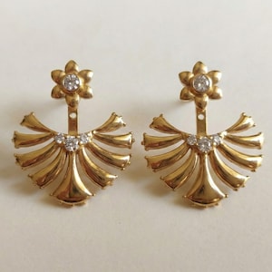 Puede incluir: Un par de pendientes dorados con un diseño floral. Cada pendiente presenta un perno en forma de flor con una piedra transparente central, una base en forma de abanico y tres piedras transparentes más pequeñas. Los pendientes se muestran sobre una superficie blanca.