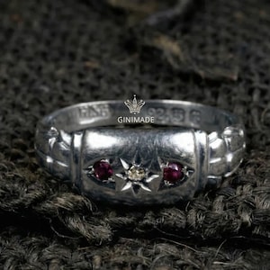 May include: A silver ring with a rectangular center section, featuring two red gemstones and a central white gemstone set within a starburst design. The ring is stamped with text and the brand name "GINIMADE".
