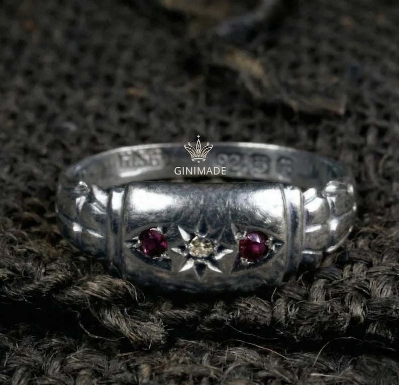 May include: A silver ring with a rectangular center section, featuring two red gemstones and a central white gemstone set within a starburst design. The ring is stamped with text and the brand name "GINIMADE".