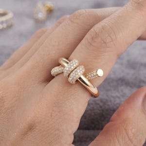 May include: A gold-toned ring featuring a knot-like design with a smooth polished band and sections encrusted with small, clear, sparkling stones. The ring wraps around the finger, showcasing a combination of plain metal and pavé-set surfaces.