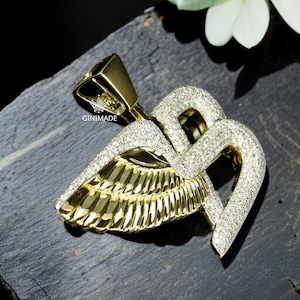 May include: A gold-colored pendant with a wing design, encrusted with small, sparkling stones. The pendant has a geometric shape at the top and is set against a dark background. The brand name "GINIMADE" is visible.