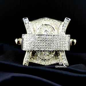 May include: A silver-coloured, ornate brooch with a rectangular centre and a shield-like design. The brooch is encrusted with numerous small, sparkling stones. The background is black, highlighting the brooch's intricate details and the text "GINIMADE".