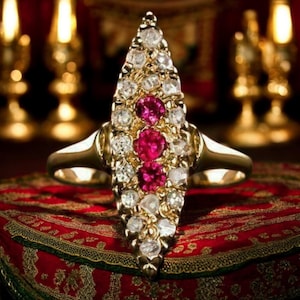 May include: A gold ring with a central, elongated design. The ring features a cluster of white diamonds and three vibrant red gemstones. The ring is set against a red surface with gold trim, with blurred background elements.