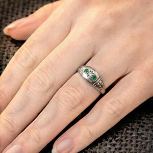 May include: A silver ring with two green gemstones and a central diamond-like stone. The ring has a decorative design on the band and is worn on a finger.