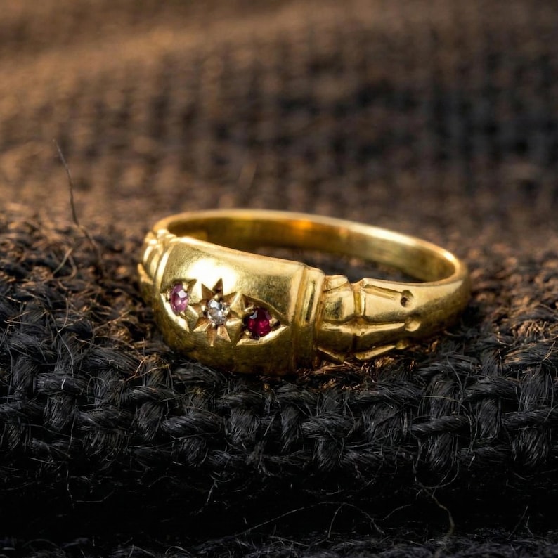 May include: A gold ring with three gemstones: two red rubies and a central diamond. The ring has a rectangular face with decorative detailing and is set against a dark, textured background.