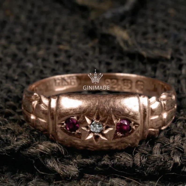 May include: Rose gold ring with a central rectangular design, flanked by two round red gemstones and a central starburst design with a small, round, clear gemstone. The ring is stamped with the text "GINIMADE".