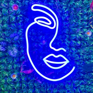 Könnte beinhalten: Ein blaues Neon-Schild mit einer weißen Umrandung eines Gesichts. Das Gesicht hat ein geschlossenes Auge, eine Nase und einen Mund.