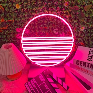 May include: A pink neon sign shaped like a sunset with a white base. The sign is sitting on a table with a black and white clapperboard and a stack of books with the text "Century Redstar" on the top book.