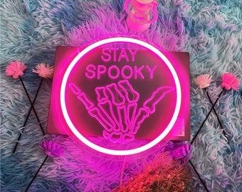 Könnte beinhalten: Rosa Neon-Schild mit einem Kreis darum. Das Schild sagt "Stay Spooky" und hat eine Skeletthand, die ein Shaka-Zeichen macht.